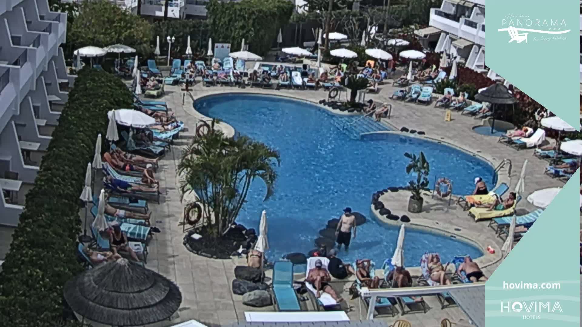 A large, kidney-shaped swimming pool surrounded by sunbathers lounging on chairs and under umbrellas on a sunny day.