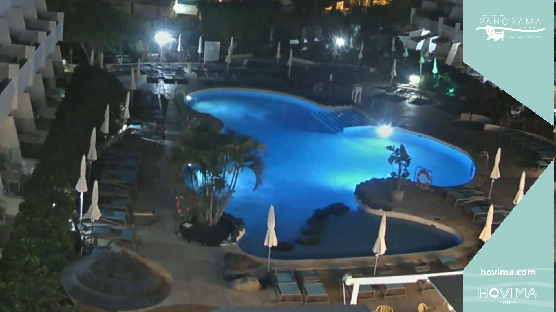 An illuminated, kidney-shaped swimming pool surrounded by lounge chairs and umbrellas at night.