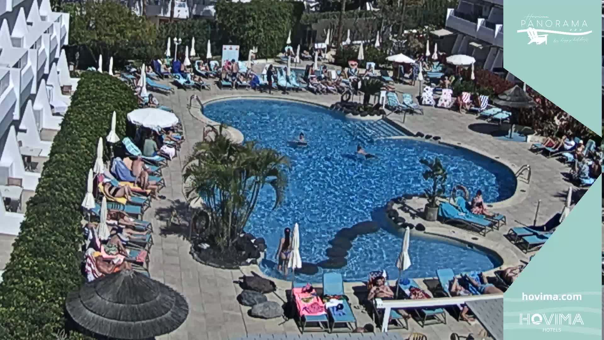 An outdoor pool with many people swimming and sunbathing on lounge chairs under umbrellas on a sunny day.