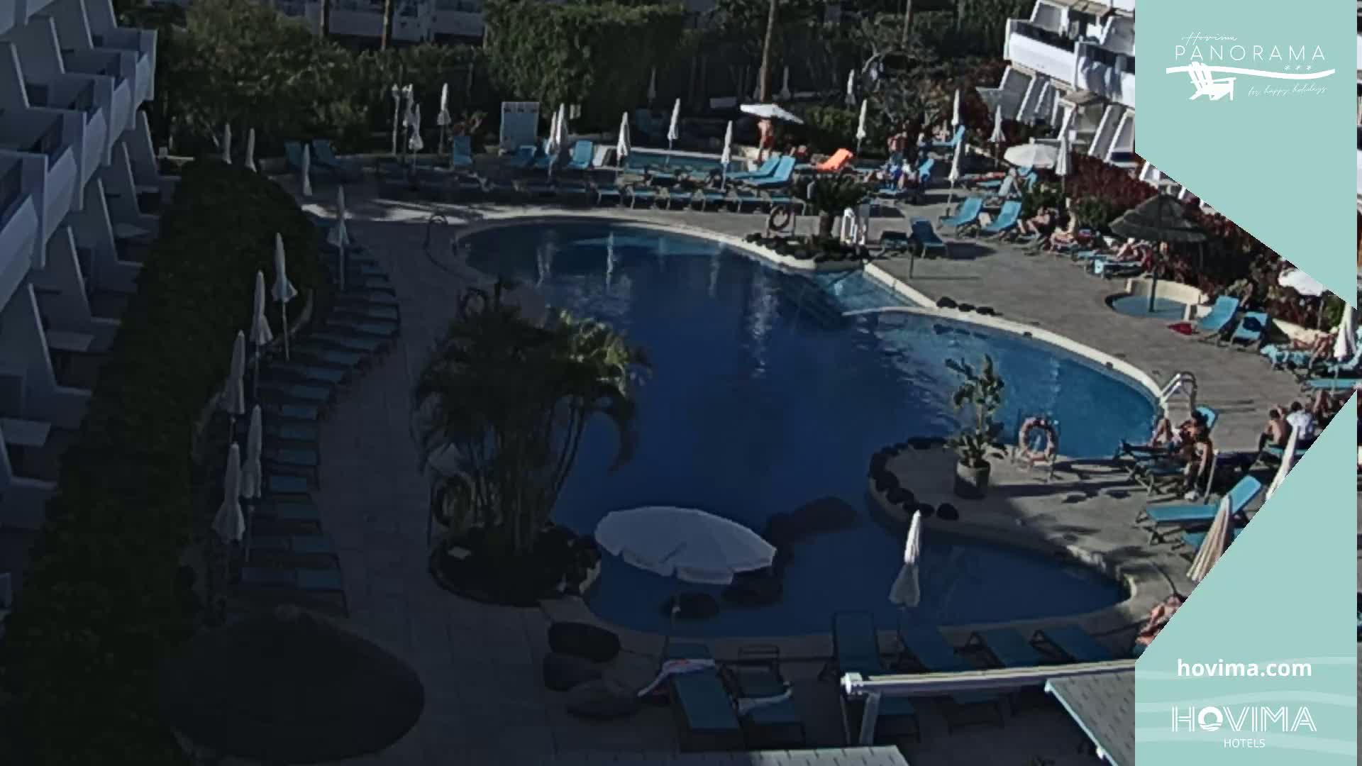 An aerial view shows a kidney-shaped swimming pool surrounded by lounge chairs and umbrellas at a resort, under sunny conditions.