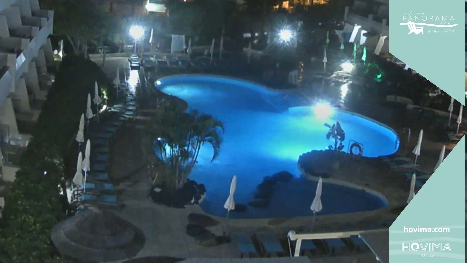 A nighttime aerial view shows a brightly lit swimming pool surrounded by lounge chairs and palm trees at a resort.