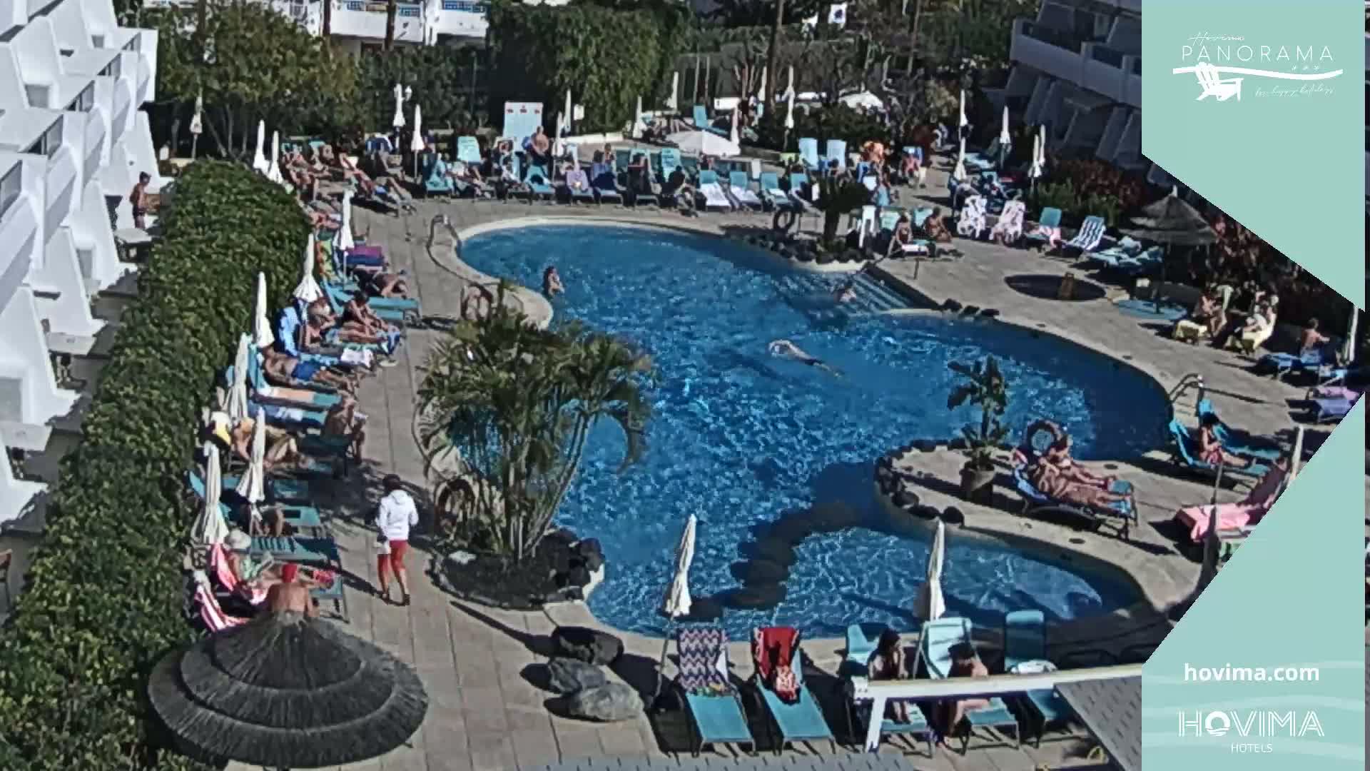 A sunny outdoor scene shows a resort pool with many people swimming and lounging on surrounding chairs and in the water.