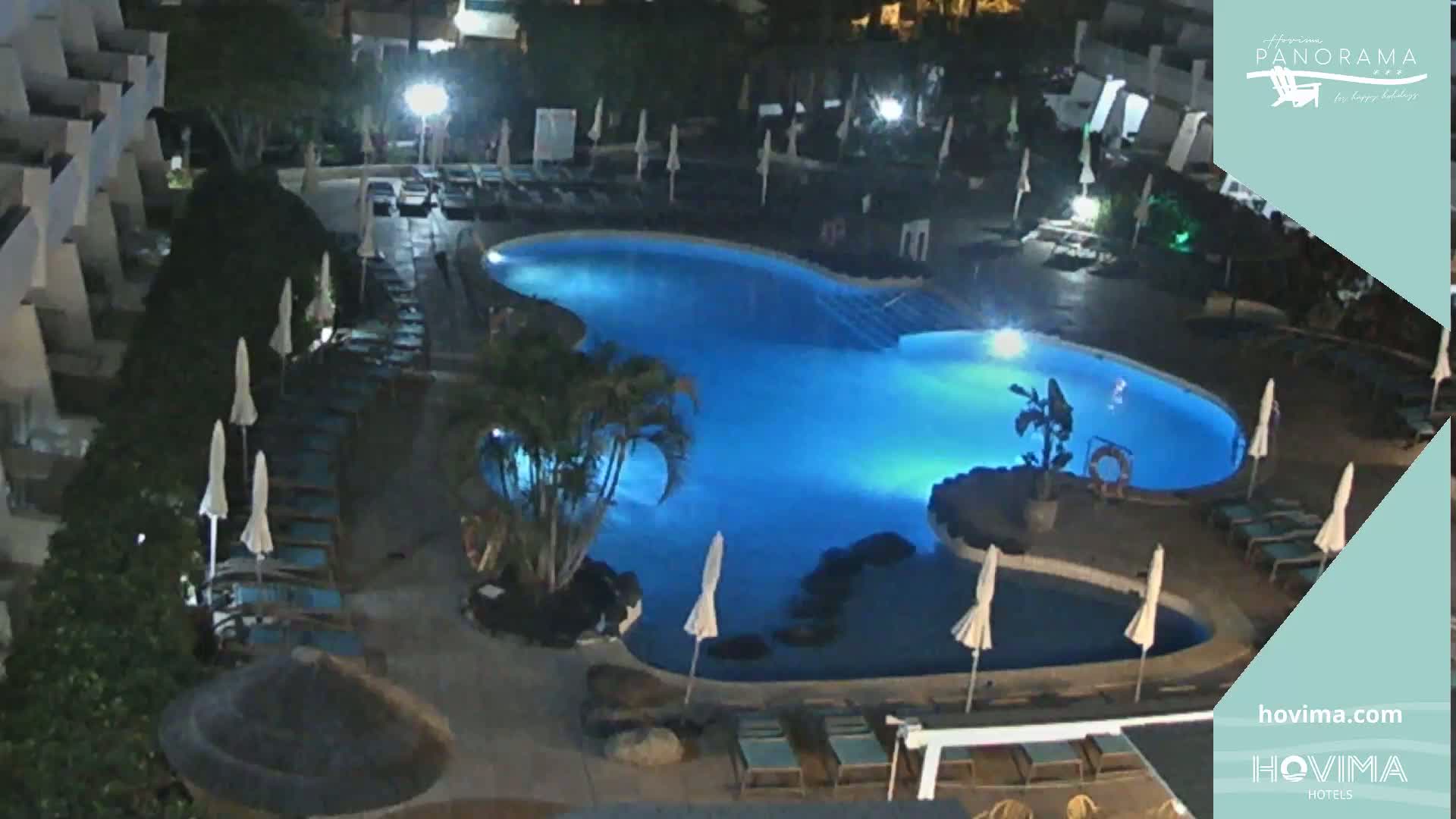 A brightly lit, kidney-shaped swimming pool at night is surrounded by lounge chairs and umbrellas in a resort setting.