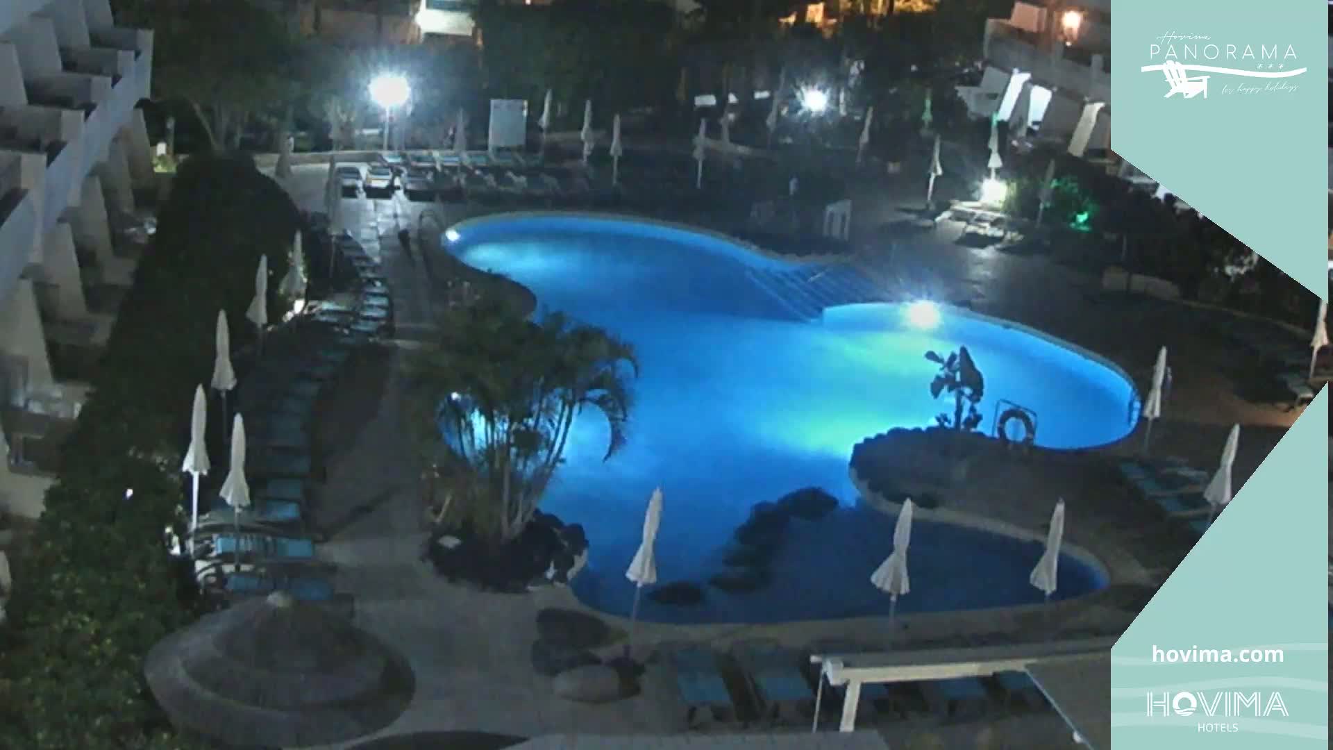 An illuminated, irregularly shaped swimming pool is surrounded by lounge chairs and umbrellas at night.