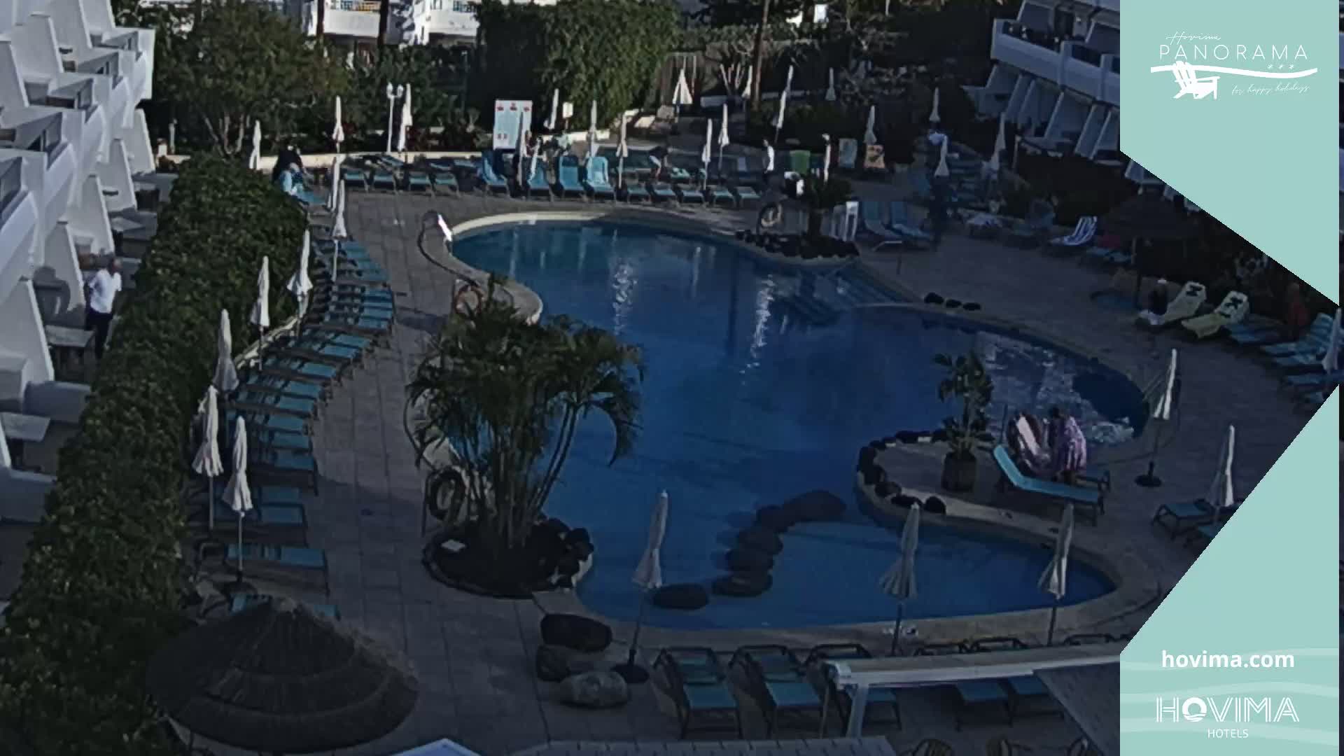 An aerial view of a resort pool area shows a curved swimming pool surrounded by lounge chairs and umbrellas under a sunny sky.