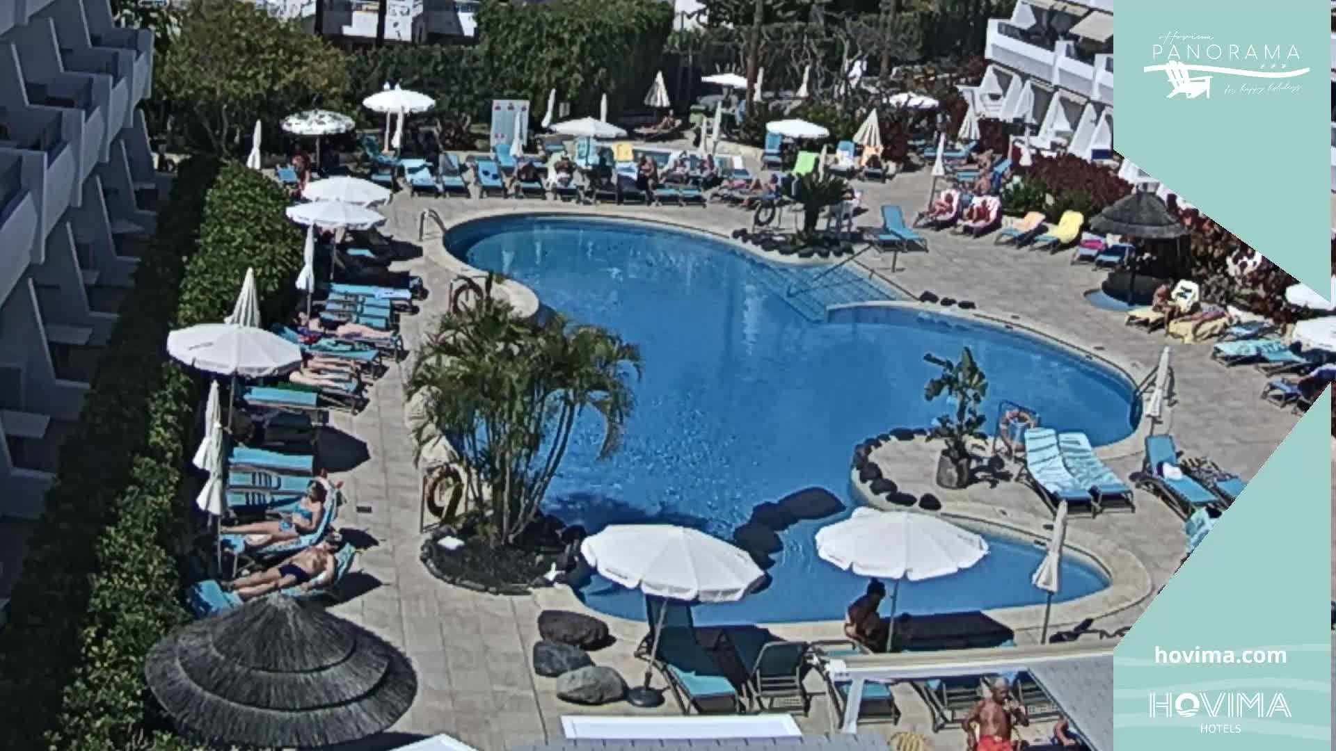 A resort pool area with many people sunbathing on lounge chairs under umbrellas on a sunny day.