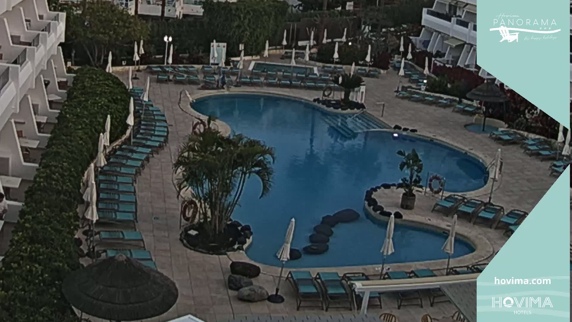 An outdoor swimming pool with surrounding lounge chairs and a building in the background, under what appears to be an overcast sky.
