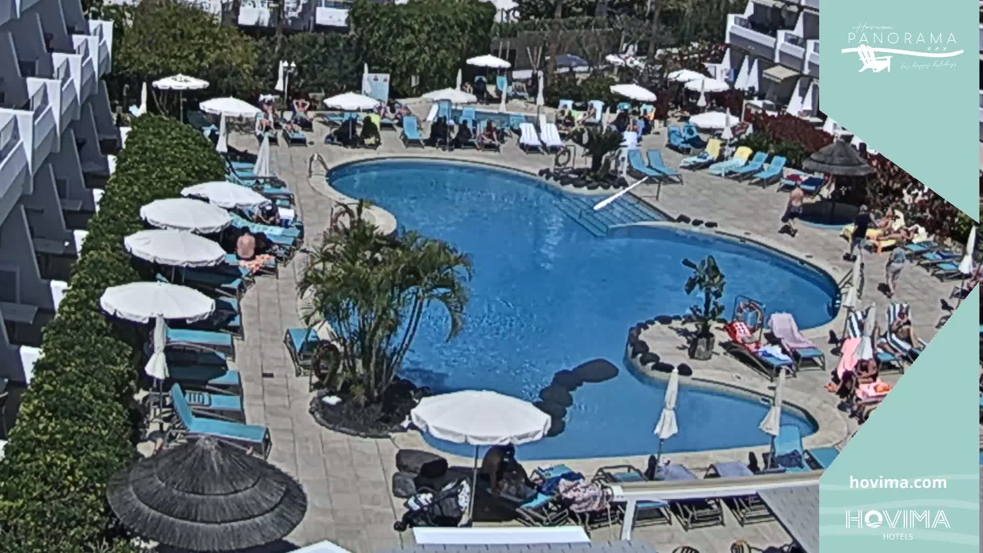 A sunny outdoor pool area with many lounge chairs and umbrellas surrounding a kidney-shaped swimming pool, populated by several people enjoying the day.