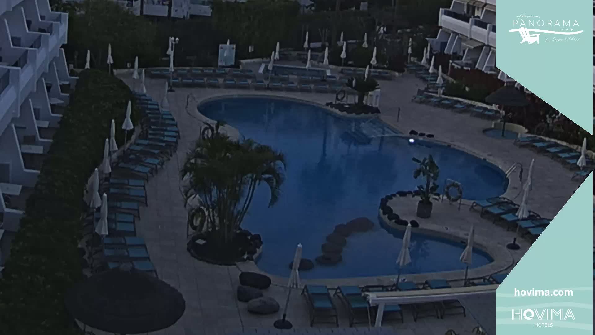 An outdoor pool area at dusk with numerous lounge chairs and umbrellas surrounding a kidney-shaped swimming pool.