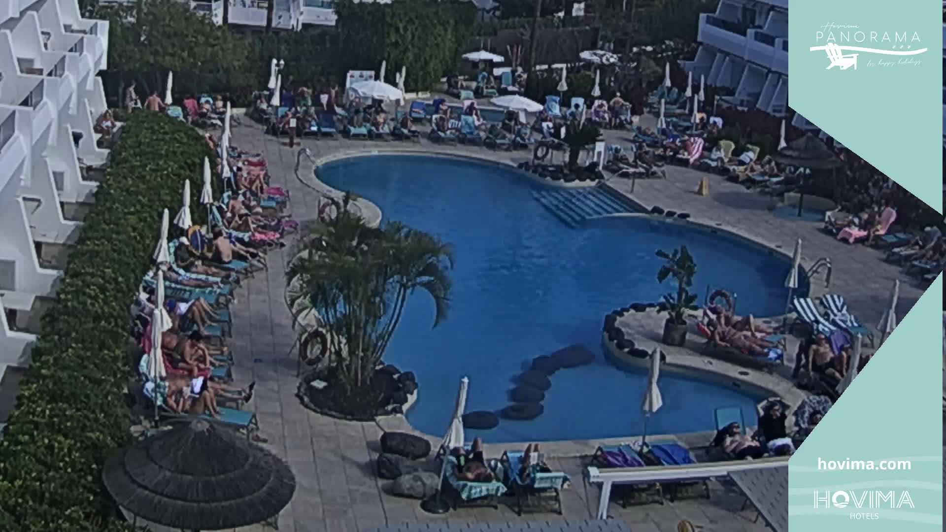 A sunny day shows a resort pool with many people lounging on deck chairs around it and several people swimming in the water.