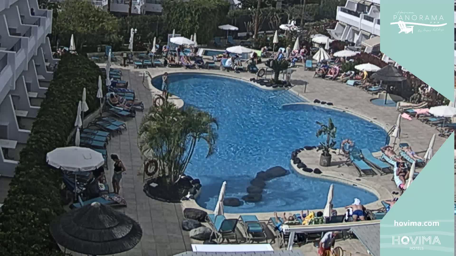 An outdoor pool area with numerous people lounging on chairs and in the water under a sunny sky.