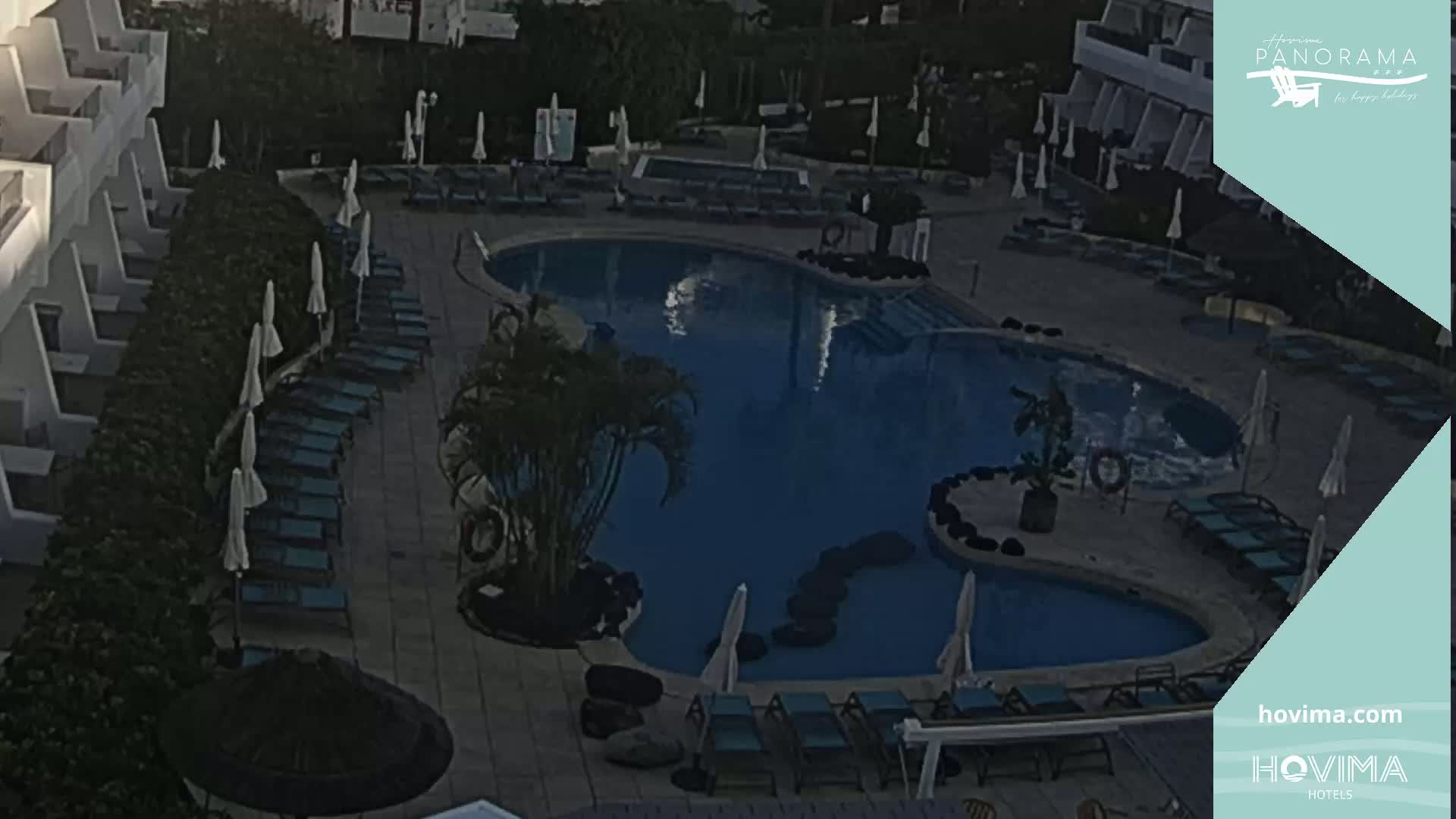 An outdoor pool area with numerous lounge chairs and umbrellas surrounding a kidney-shaped pool, under what appears to be an evening sky.