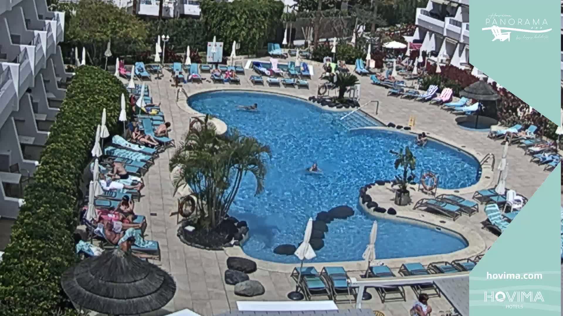 An outdoor swimming pool with many people lounging on surrounding chairs and in the water on a sunny day.