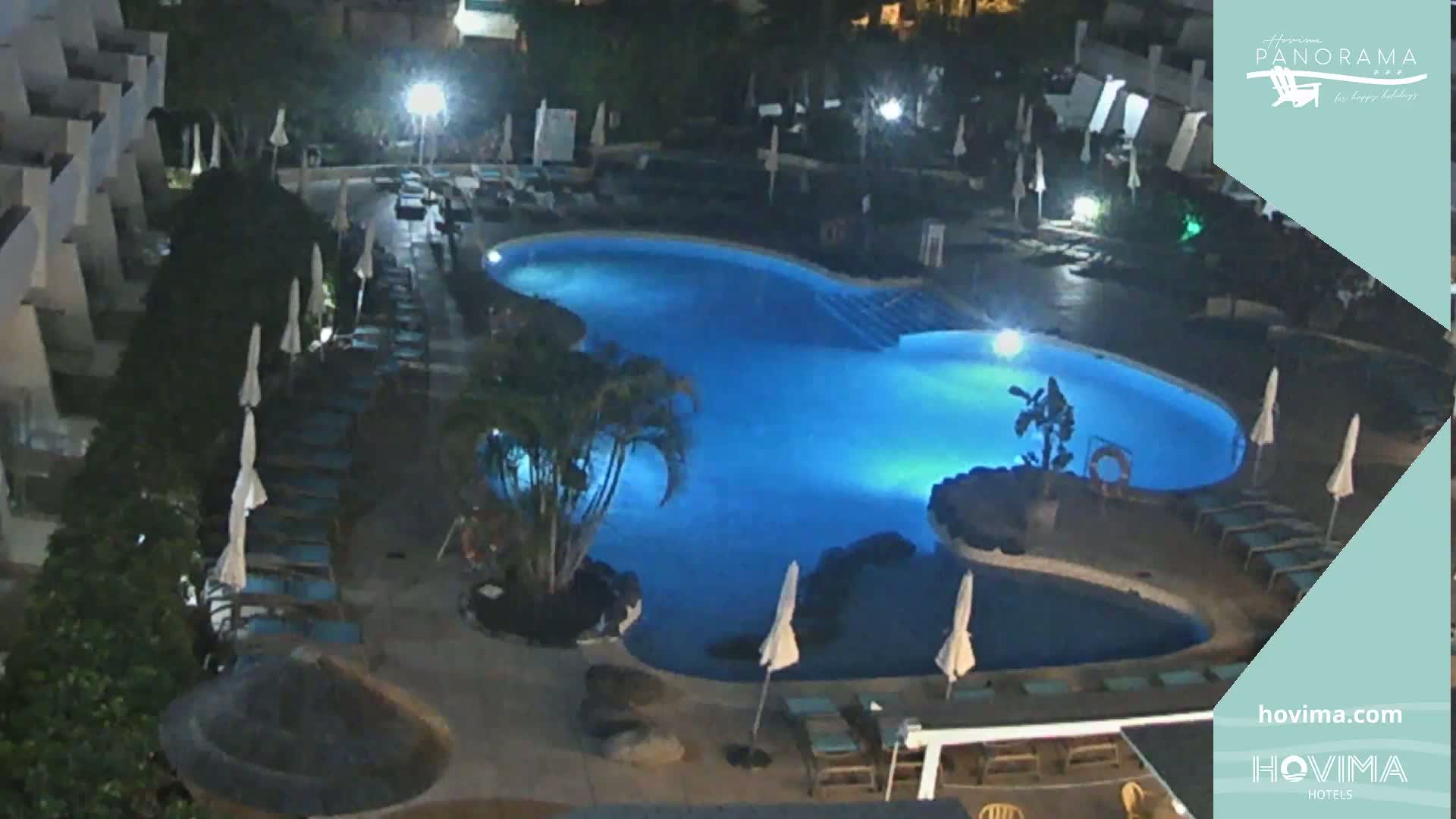 An illuminated, kidney-shaped swimming pool at night is surrounded by lounge chairs and umbrellas, nestled within a resort complex under dark skies.