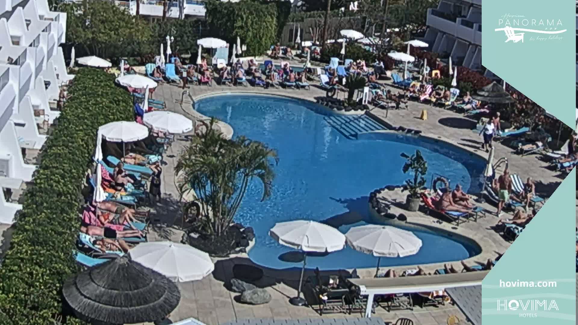 A sunny outdoor scene shows a resort pool area with many people relaxing on lounge chairs and in the water under numerous umbrellas.