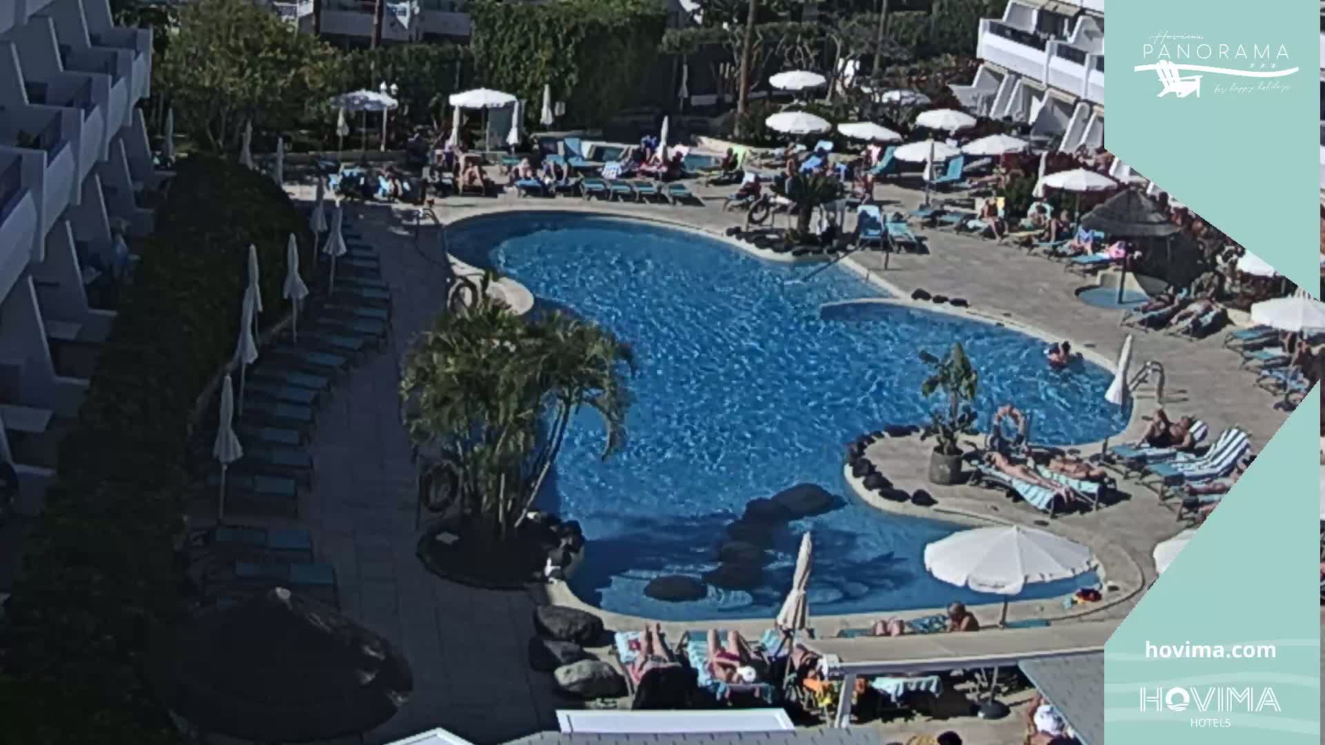 A sunny outdoor scene shows a large swimming pool surrounded by lounge chairs and umbrellas, with many people relaxing and swimming on a sunny day.