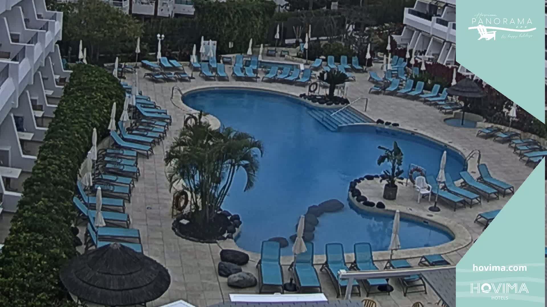 An outdoor pool with numerous blue lounge chairs surrounding it, situated next to a multi-story building under an overcast sky.