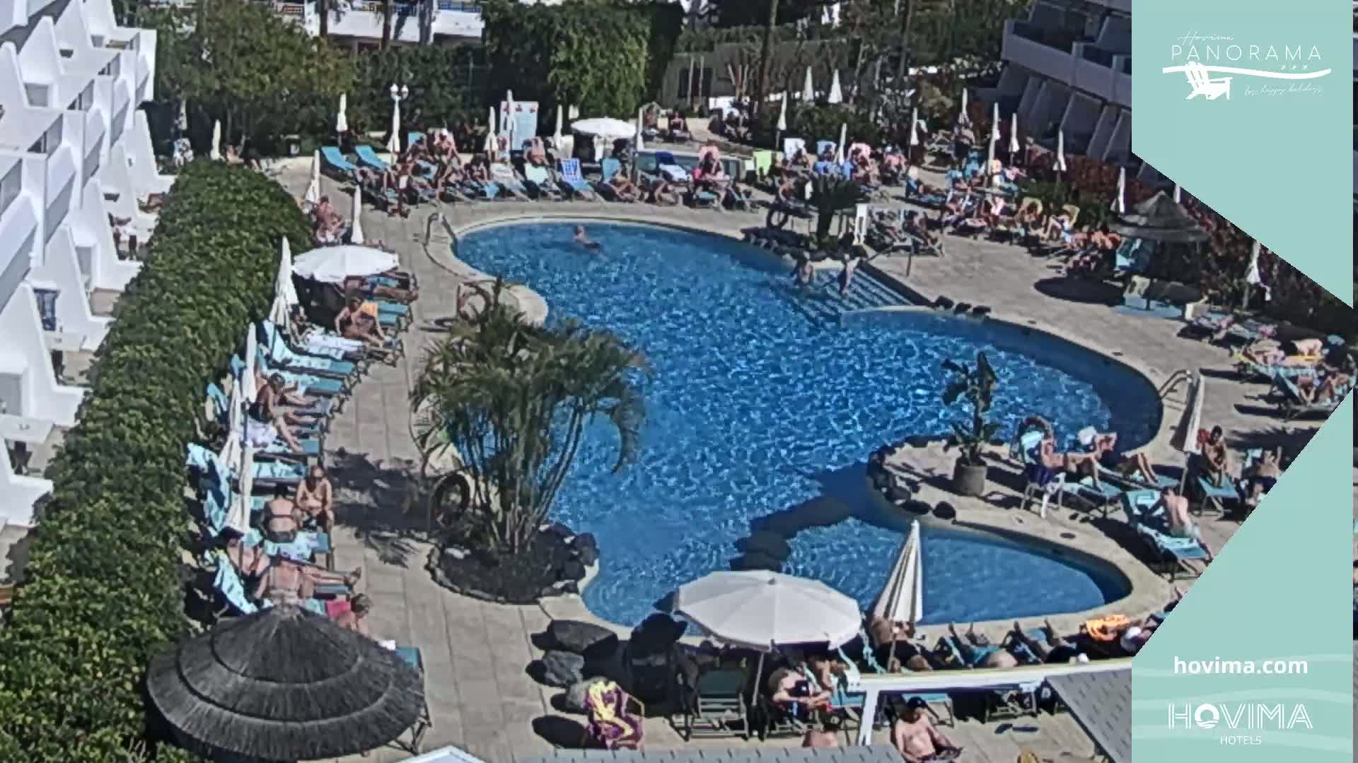 An aerial view shows a sunny resort pool area with numerous people lounging by the pool and swimming.