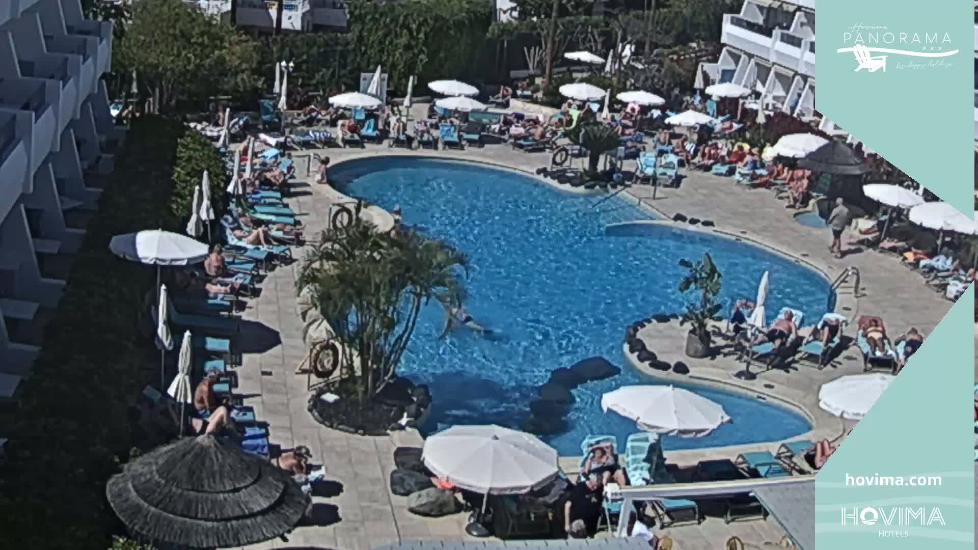 A sunny outdoor scene shows a large, winding swimming pool surrounded by people lounging on chairs and under umbrellas near a building.