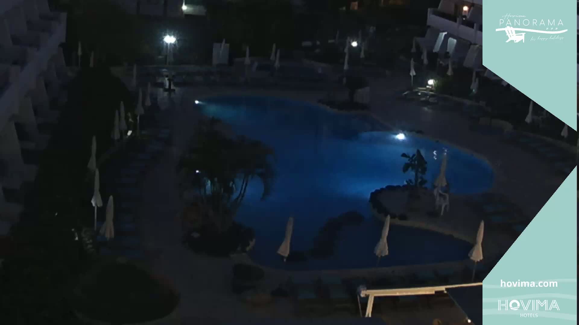 A nighttime aerial view shows a dark blue illuminated swimming pool surrounded by closed umbrellas and dark lounge chairs.