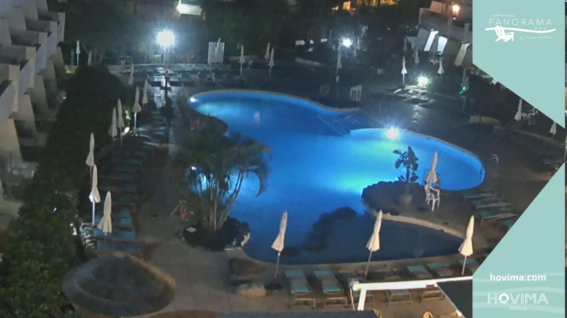 A nighttime aerial view of a resort pool area shows a lit, kidney-shaped swimming pool surrounded by lounge chairs and umbrellas.