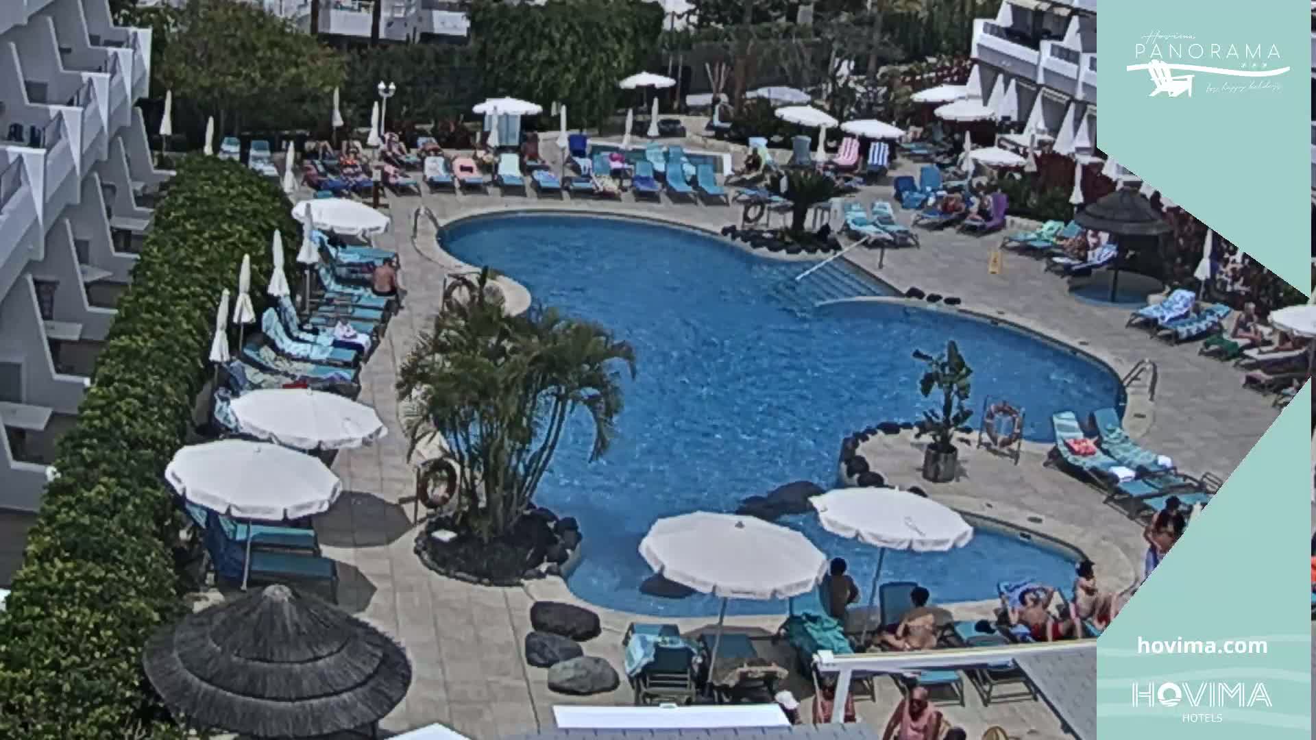 An outdoor swimming pool surrounded by lounge chairs, umbrellas, and a building, on a sunny day.