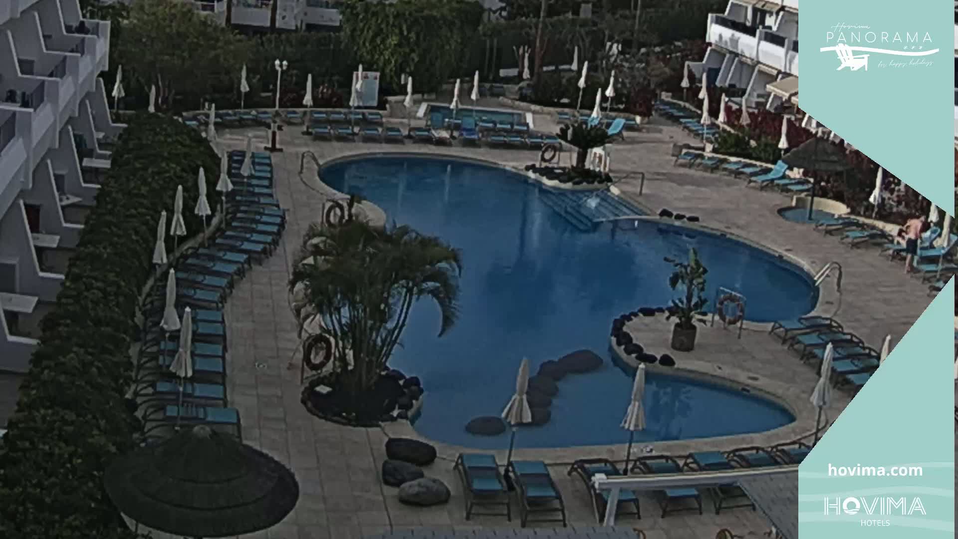 An aerial view of a resort pool area shows a kidney-shaped pool surrounded by lounge chairs and umbrellas under a partly cloudy sky.