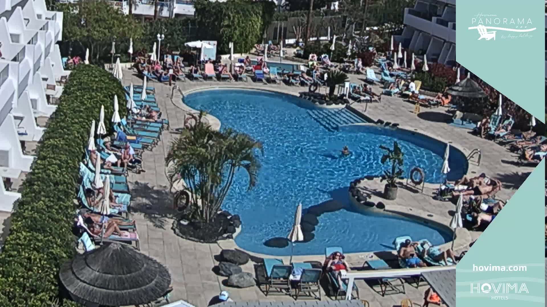 An outdoor pool surrounded by sunbathers and lounge chairs under umbrellas on a sunny day.