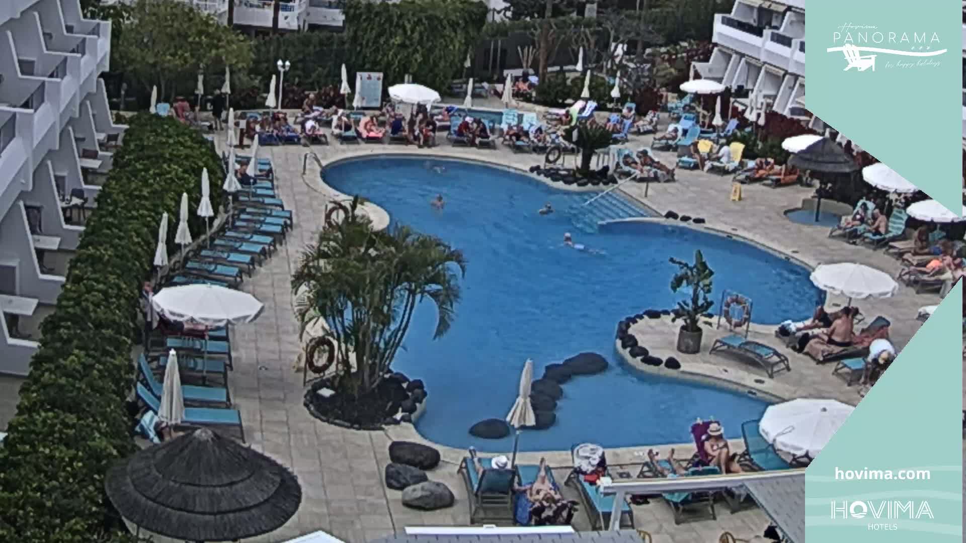 An outdoor swimming pool surrounded by lounge chairs and umbrellas, with people relaxing and swimming on a sunny day.