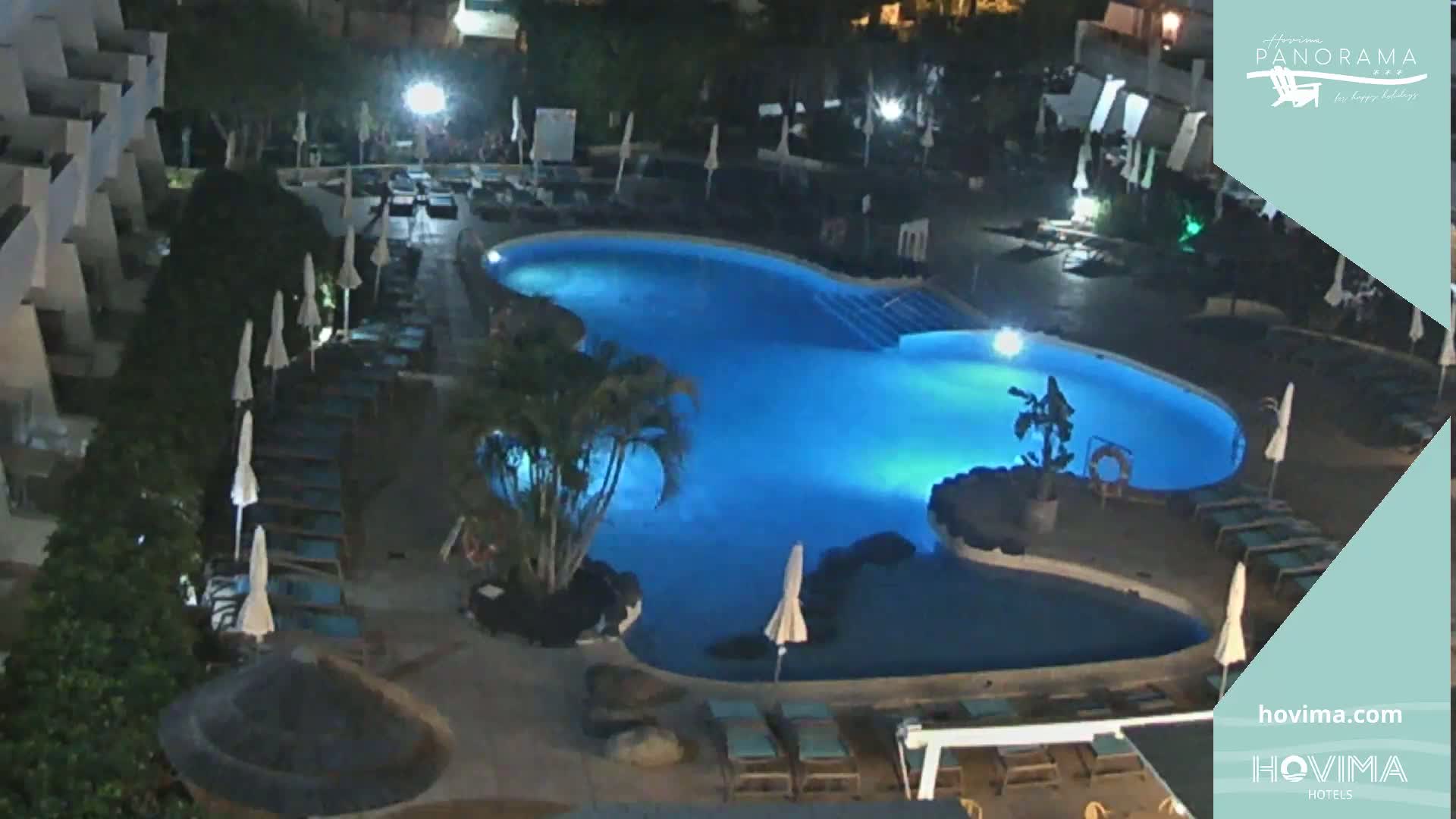 An illuminated, kidney-shaped swimming pool at night, surrounded by lounge chairs and umbrellas, next to a building.