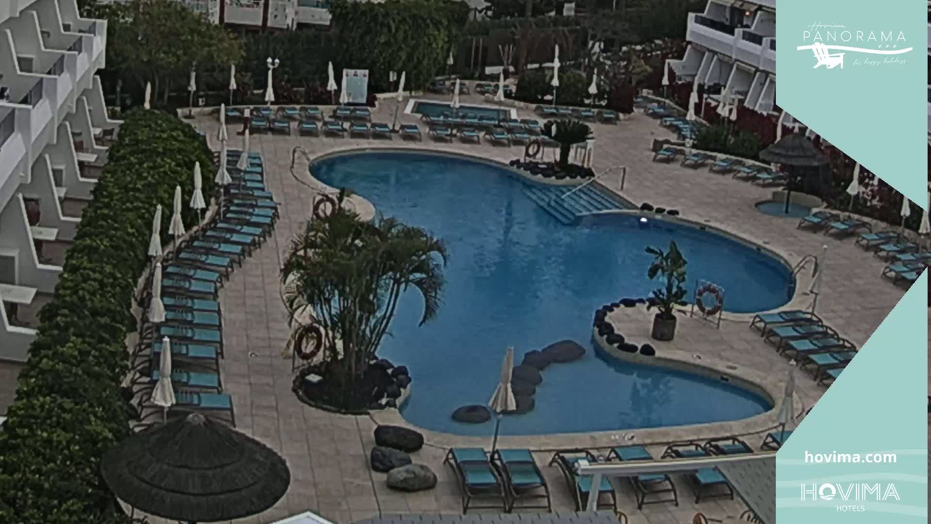 An overhead view shows a kidney-shaped swimming pool surrounded by lounge chairs and umbrellas at a resort, with overcast skies.
