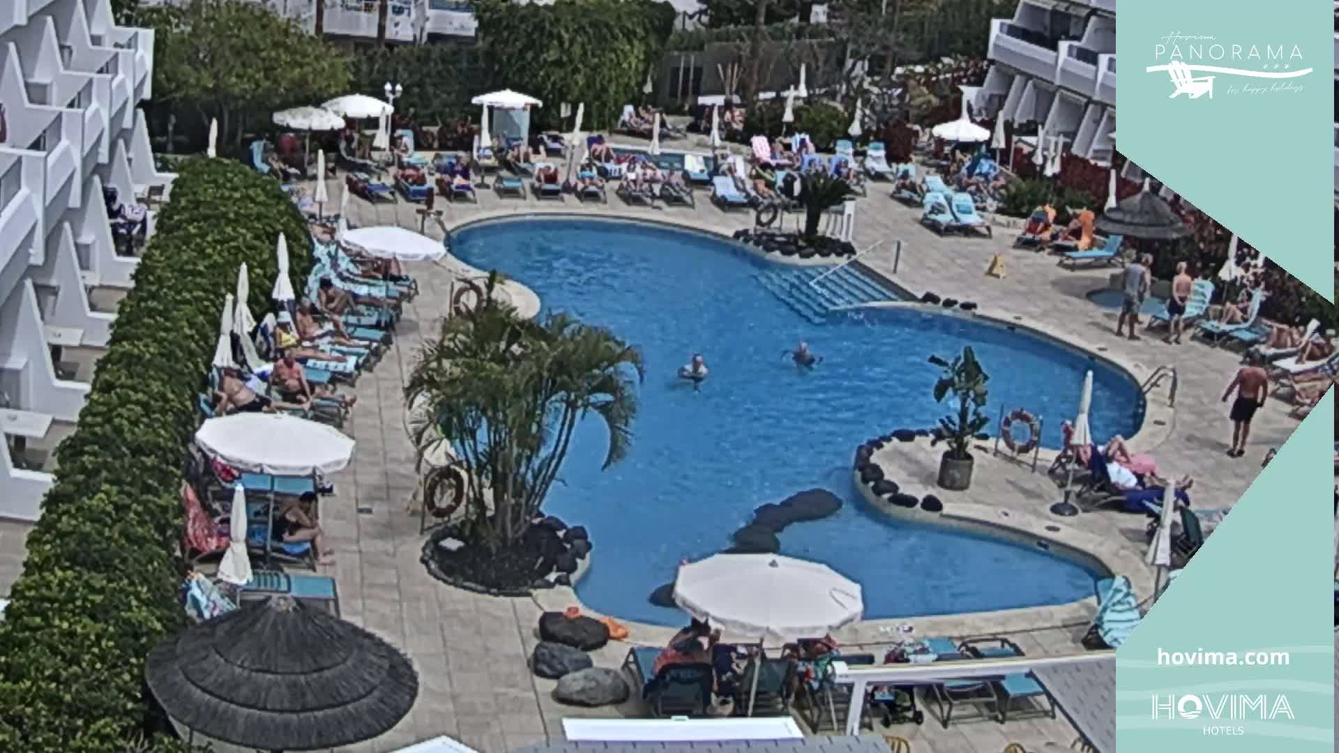 A resort pool area with many people lounging around the pool and on the surrounding deck chairs under sunny skies.