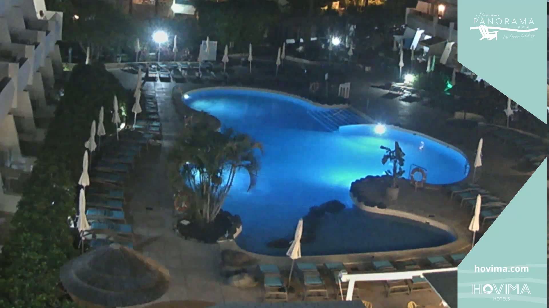 A nighttime view of an illuminated kidney-shaped swimming pool surrounded by lounge chairs and umbrellas at a resort.