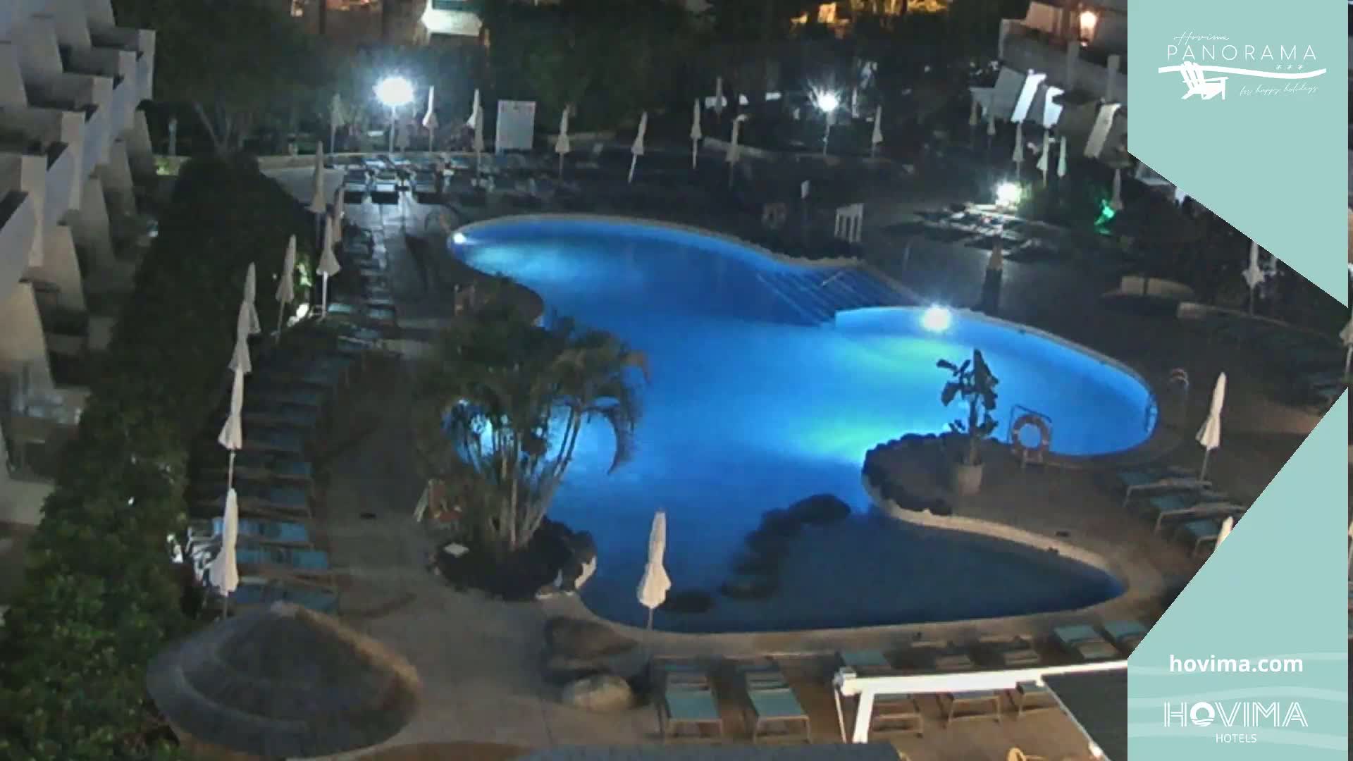 A nighttime view of a brightly lit swimming pool surrounded by lounge chairs and palm trees at a resort.