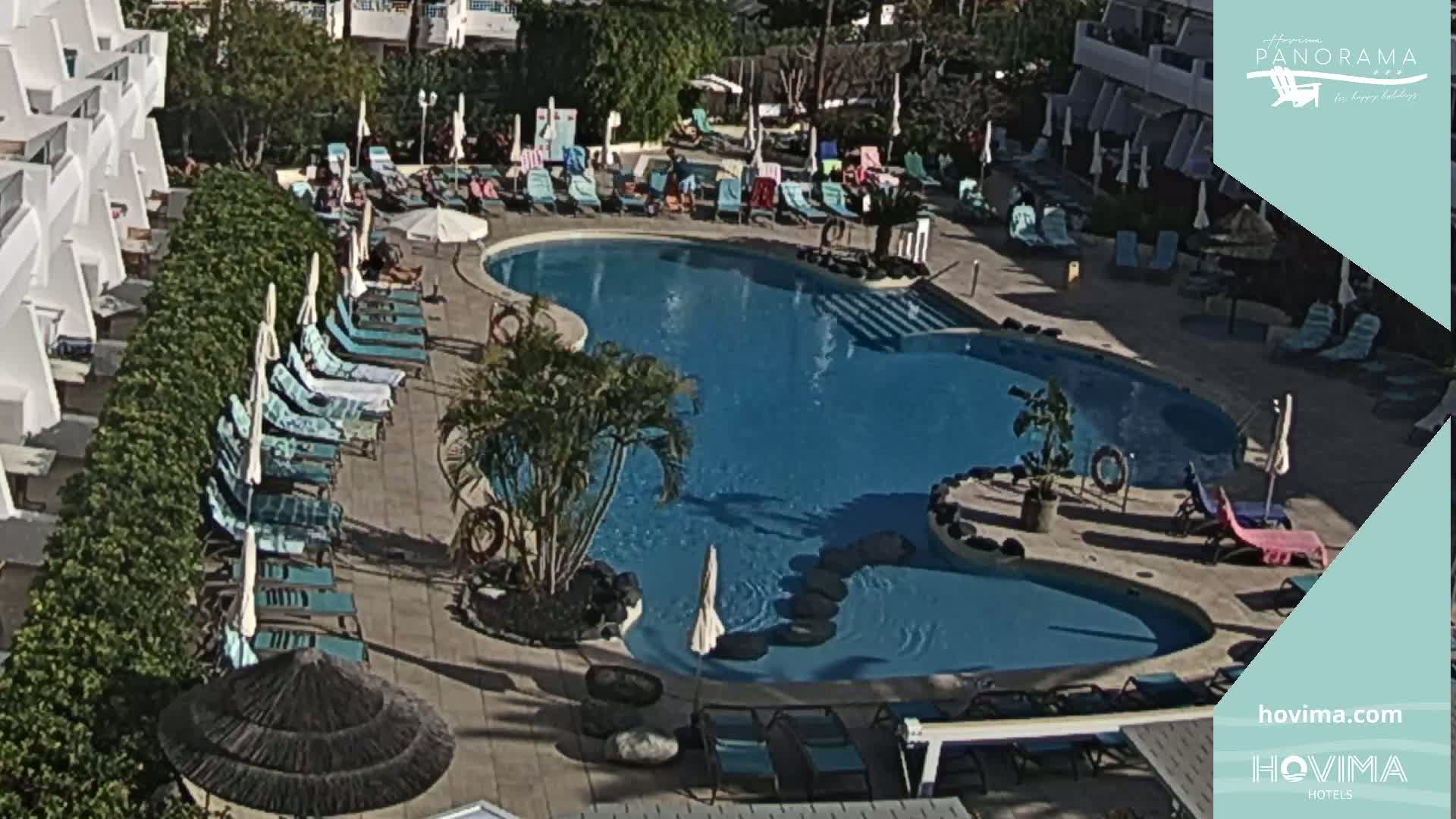 An aerial view of a resort pool area on a sunny day shows a kidney-shaped swimming pool surrounded by lounge chairs and umbrellas.