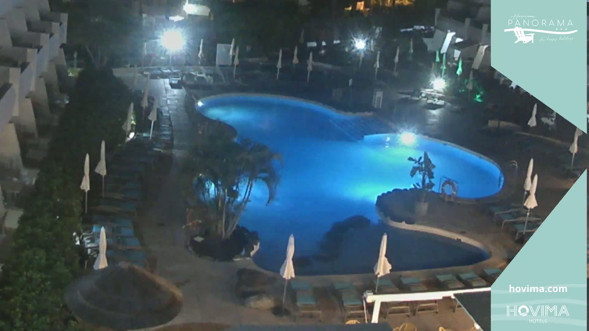An illuminated swimming pool at night, surrounded by lounge chairs and umbrellas, is situated within a resort.