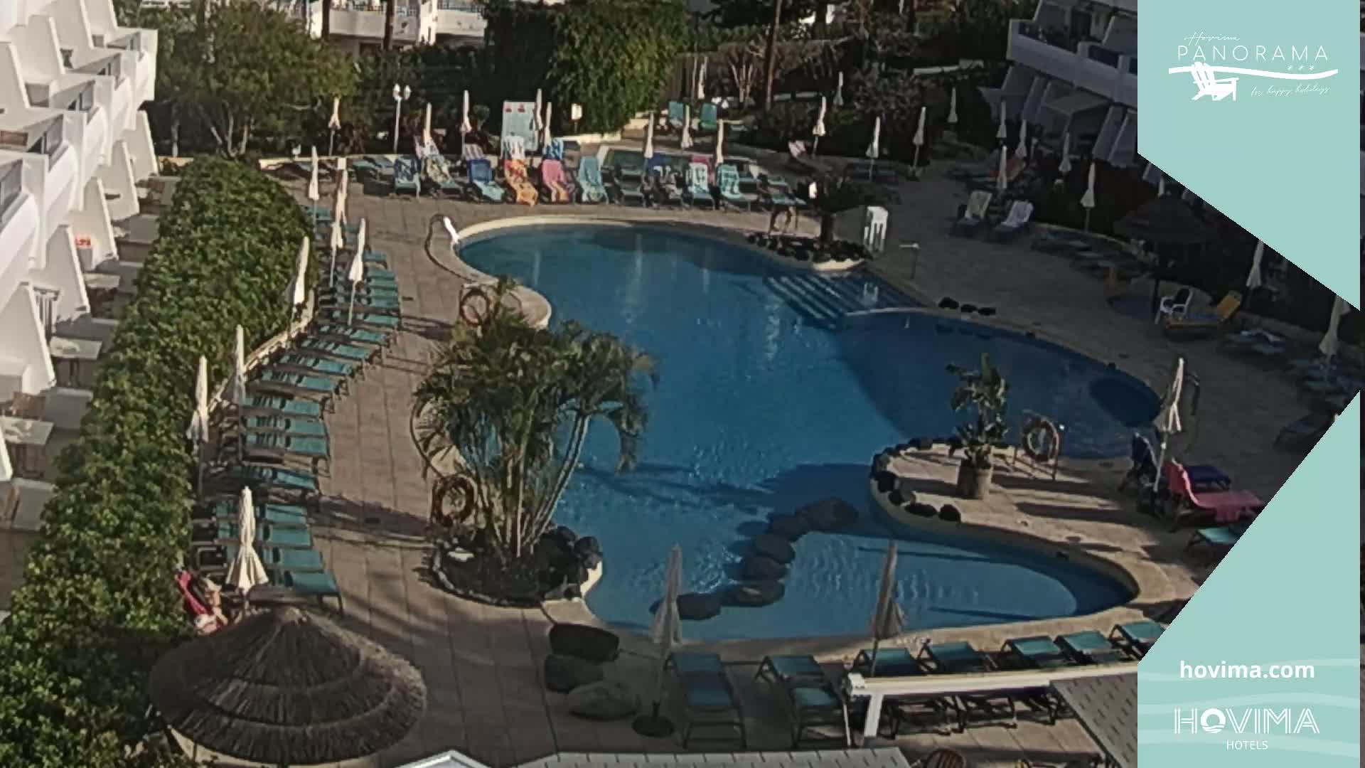 A kidney-shaped swimming pool surrounded by lounge chairs and umbrellas sits next to a white building on a sunny day.