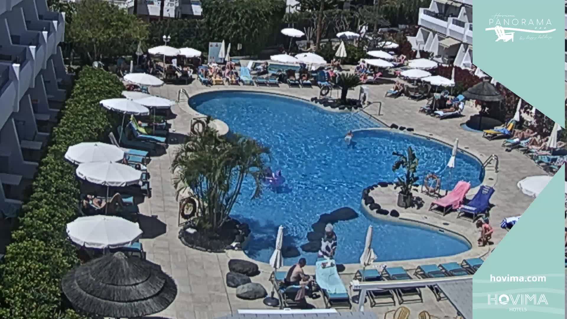 A sunny outdoor scene shows a resort pool with people swimming and lounging on chairs under umbrellas.