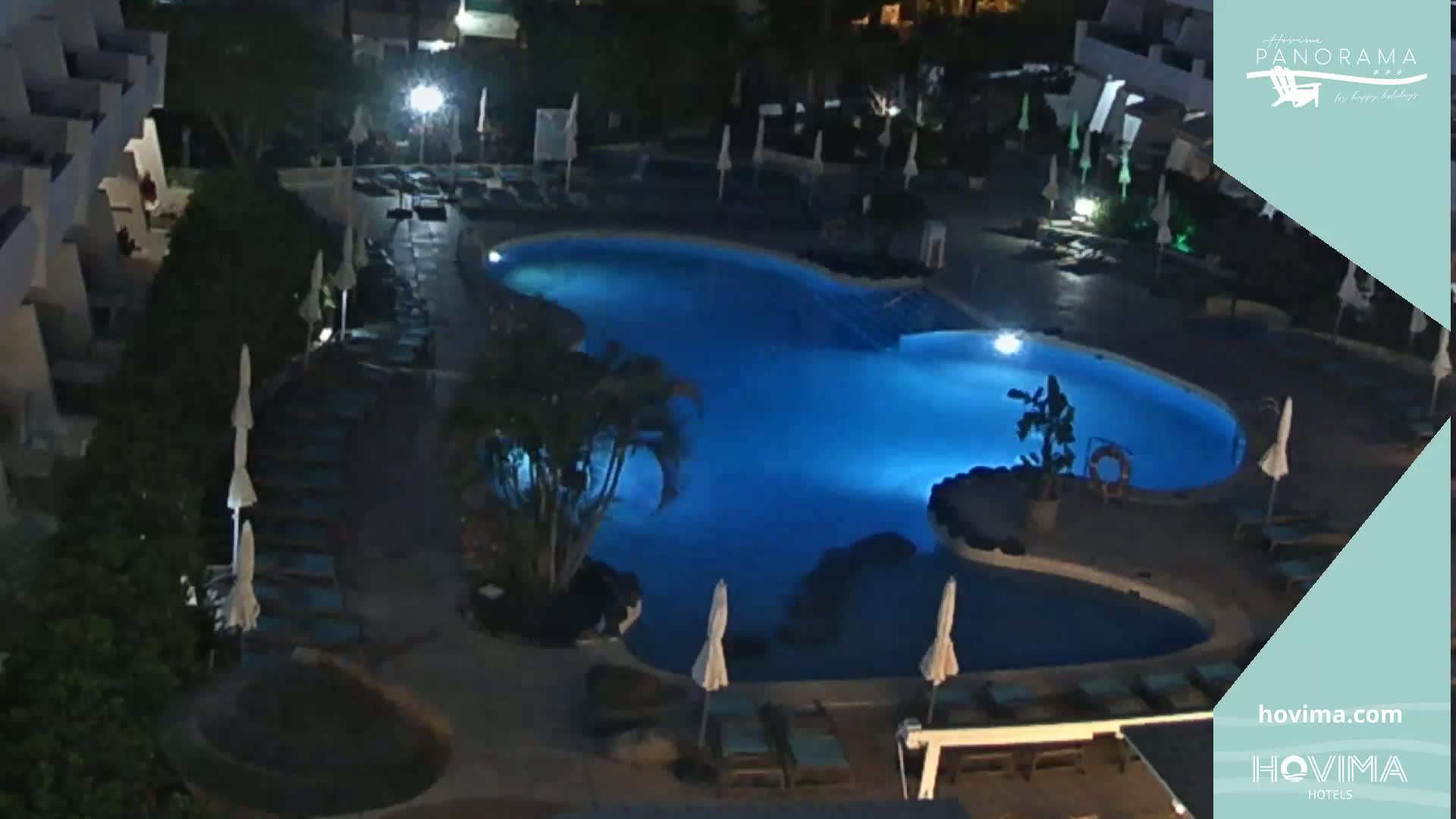 A nighttime aerial view of a resort pool area with closed umbrellas surrounding a lit pool.