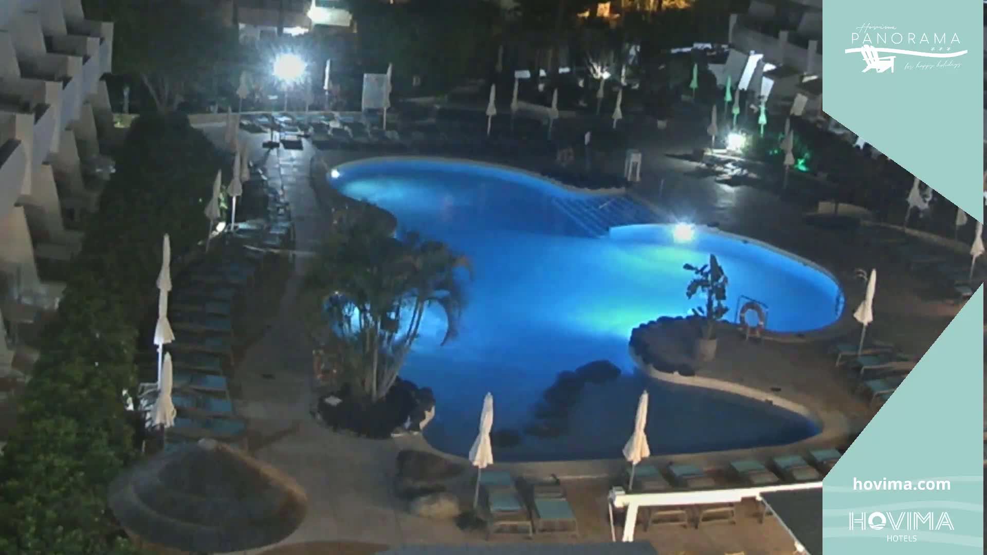 An illuminated, kidney-shaped swimming pool at night, surrounded by lounge chairs and umbrellas, sits within a resort complex.