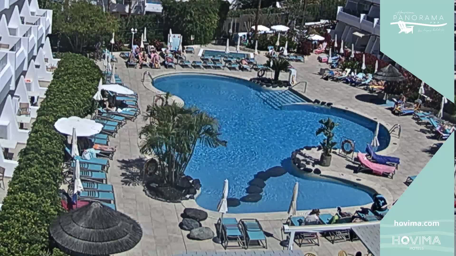 An aerial view shows a resort pool area with sunbathers lounging by a kidney-shaped pool on a sunny day.