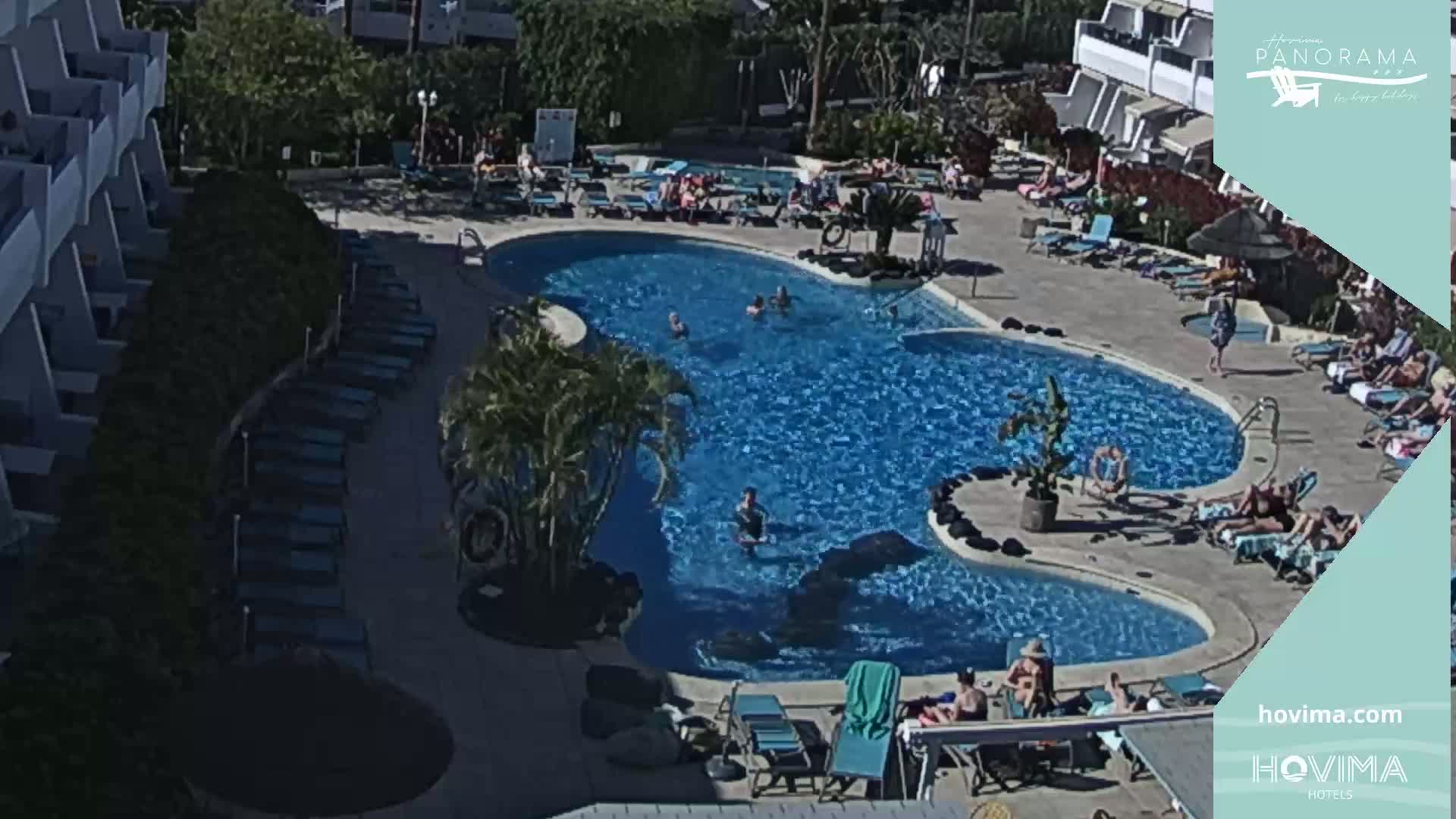 An outdoor swimming pool with people swimming and lounging on surrounding chairs and sunbeds under sunny skies.