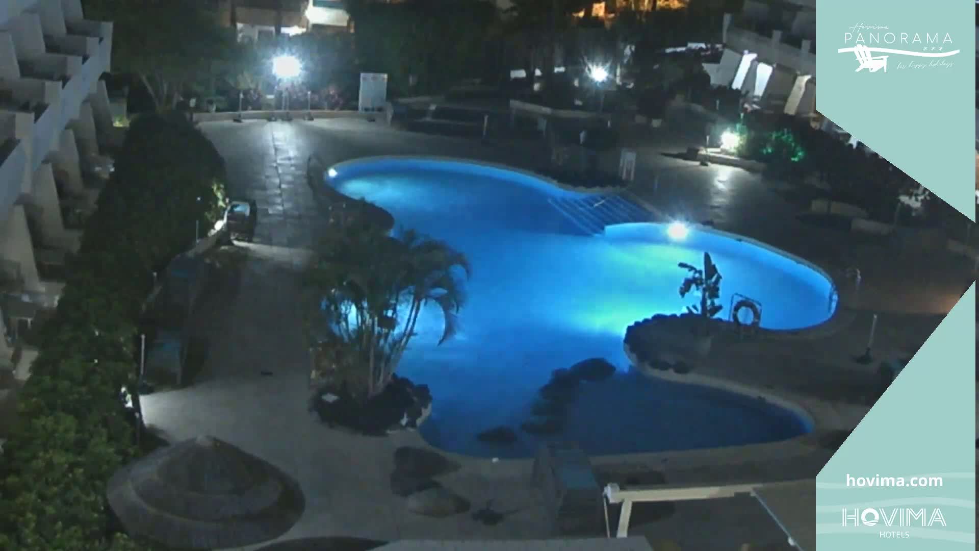 A nighttime aerial view shows a kidney-shaped swimming pool illuminated in blue, surrounded by palm trees and building structures.