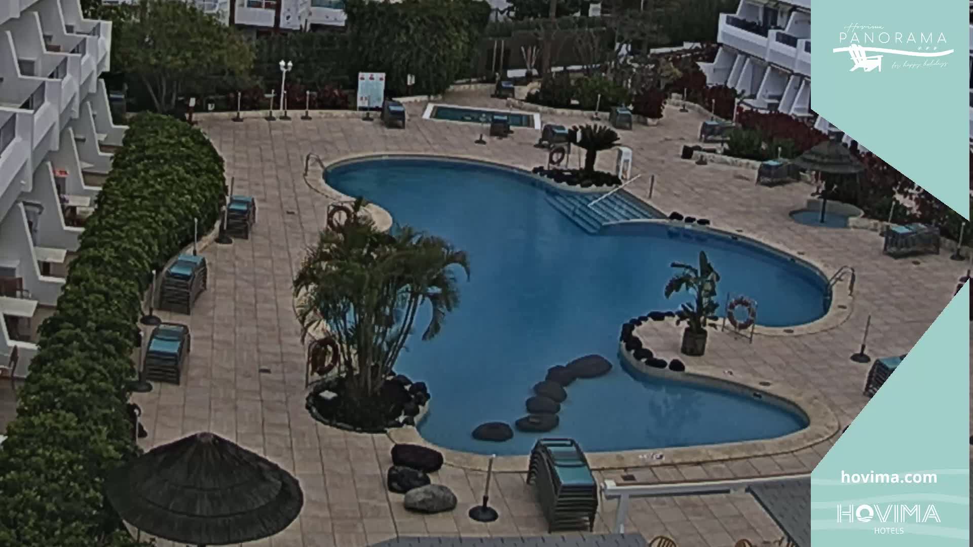 An outdoor pool with a curved shape, surrounded by lounge chairs, potted plants, and buildings on an overcast day.