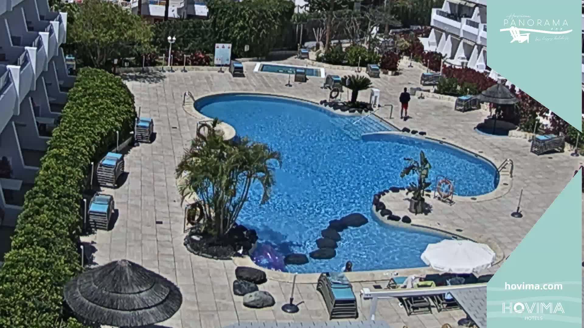 An aerial view shows a swimming pool with a surrounding patio area, several lounge chairs, and umbrellas under a sunny sky.