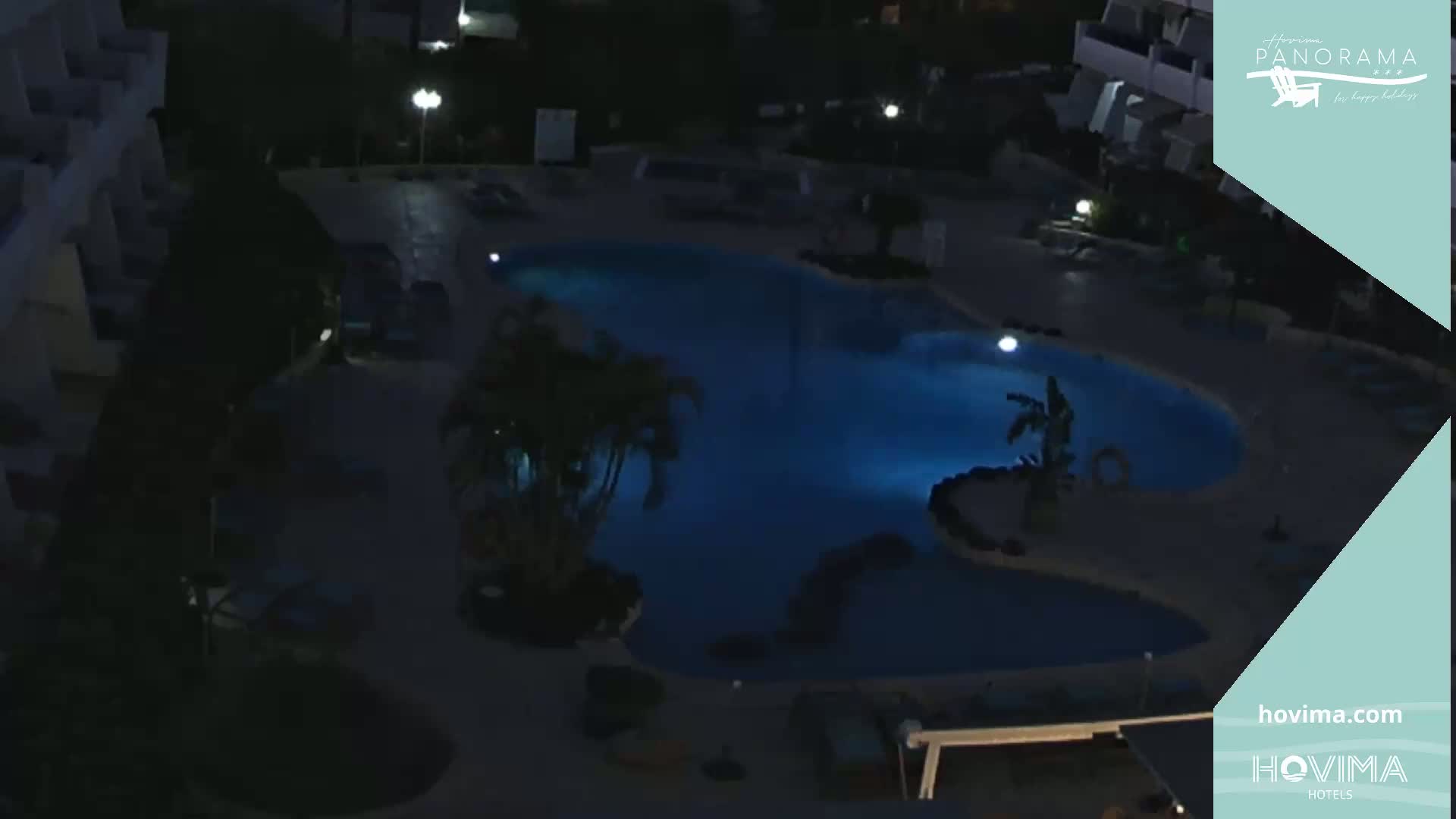 An illuminated swimming pool at night is surrounded by dark buildings and palm trees.