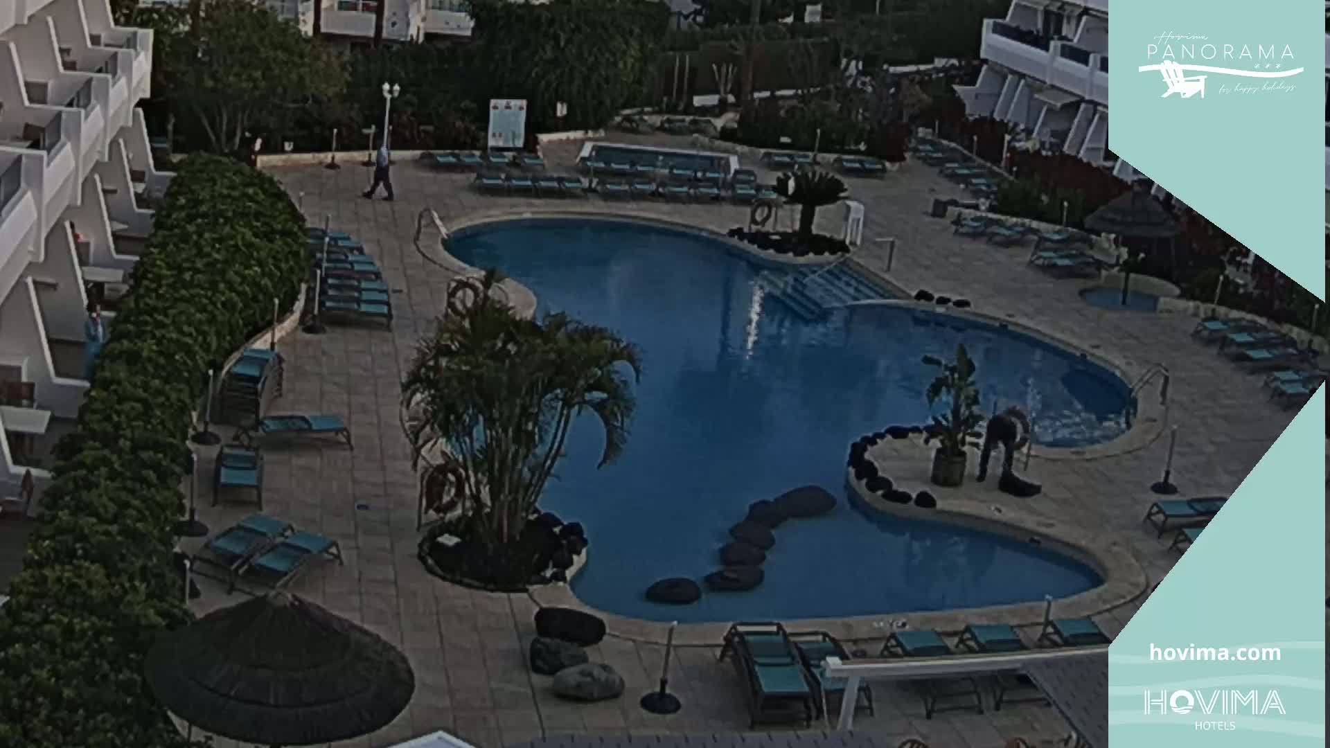 An aerial view shows a resort pool area with numerous empty lounge chairs surrounding a kidney-shaped pool under a partly cloudy sky.