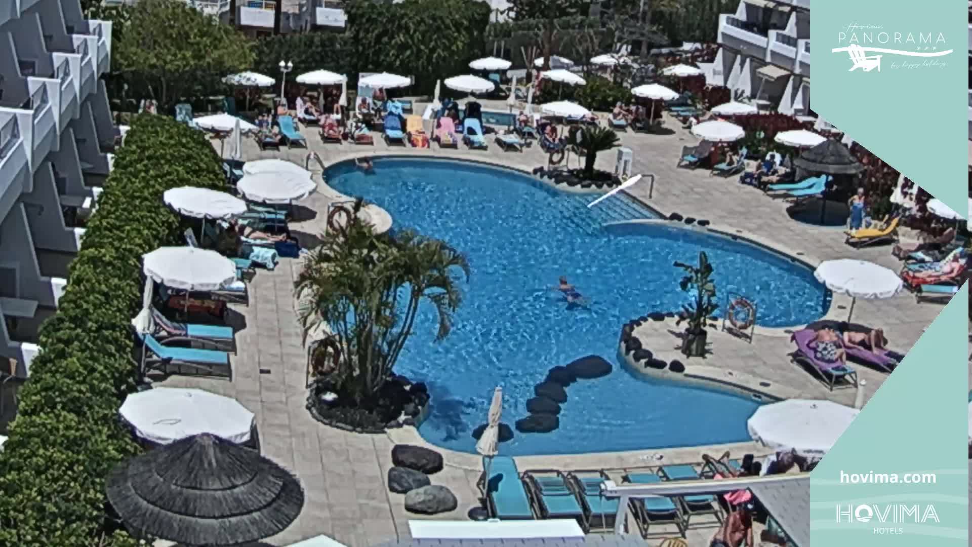 An outdoor pool surrounded by lounge chairs and umbrellas, with people swimming and sunbathing under a sunny sky.
