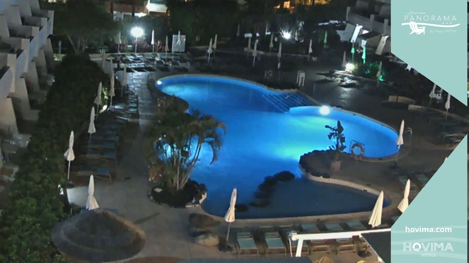 A nighttime aerial view of a resort pool area with surrounding lounge chairs and umbrellas, illuminated by artificial lighting.