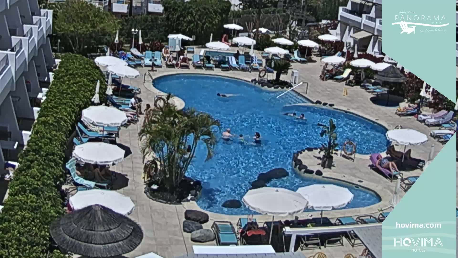An outdoor pool surrounded by lounge chairs, umbrellas, and a building, under sunny skies.